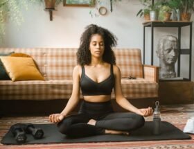 A woman meditating in a seated position on a yoga mat, surrounded by plants and fitness equipment in a cozy living room