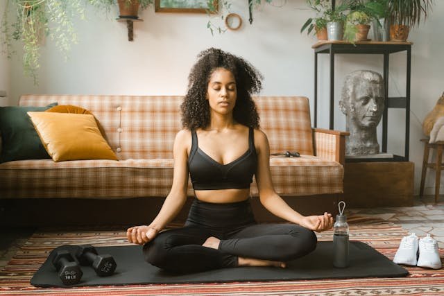 A woman meditating in a seated position on a yoga mat, surrounded by plants and fitness equipment in a cozy living room