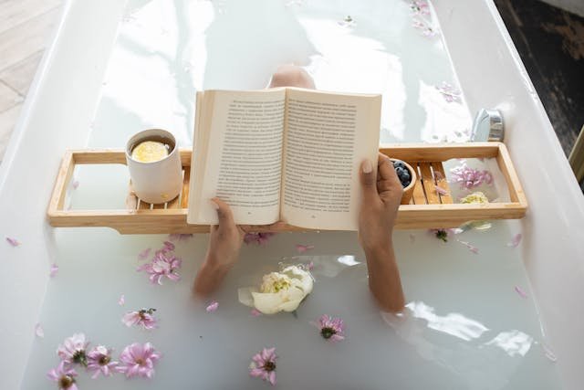 A person relaxes and reads a book in a bathtub filled with water and floating flower petals, creating a peaceful wellness space