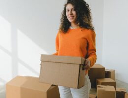 A woman wearing an orange sweater and white pants, exemplifying versatile pieces for your moving day outfit, holds a cardboard box in a room filled with more boxes