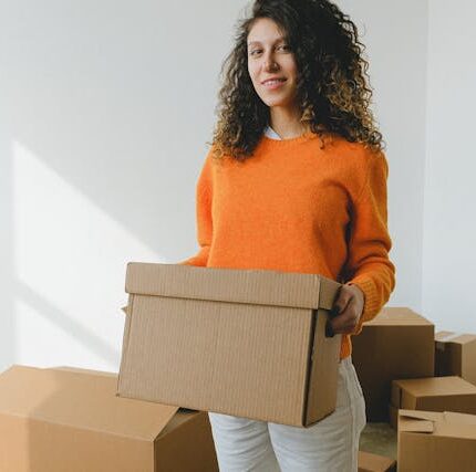 A woman wearing an orange sweater and white pants, exemplifying versatile pieces for your moving day outfit, holds a cardboard box in a room filled with more boxes